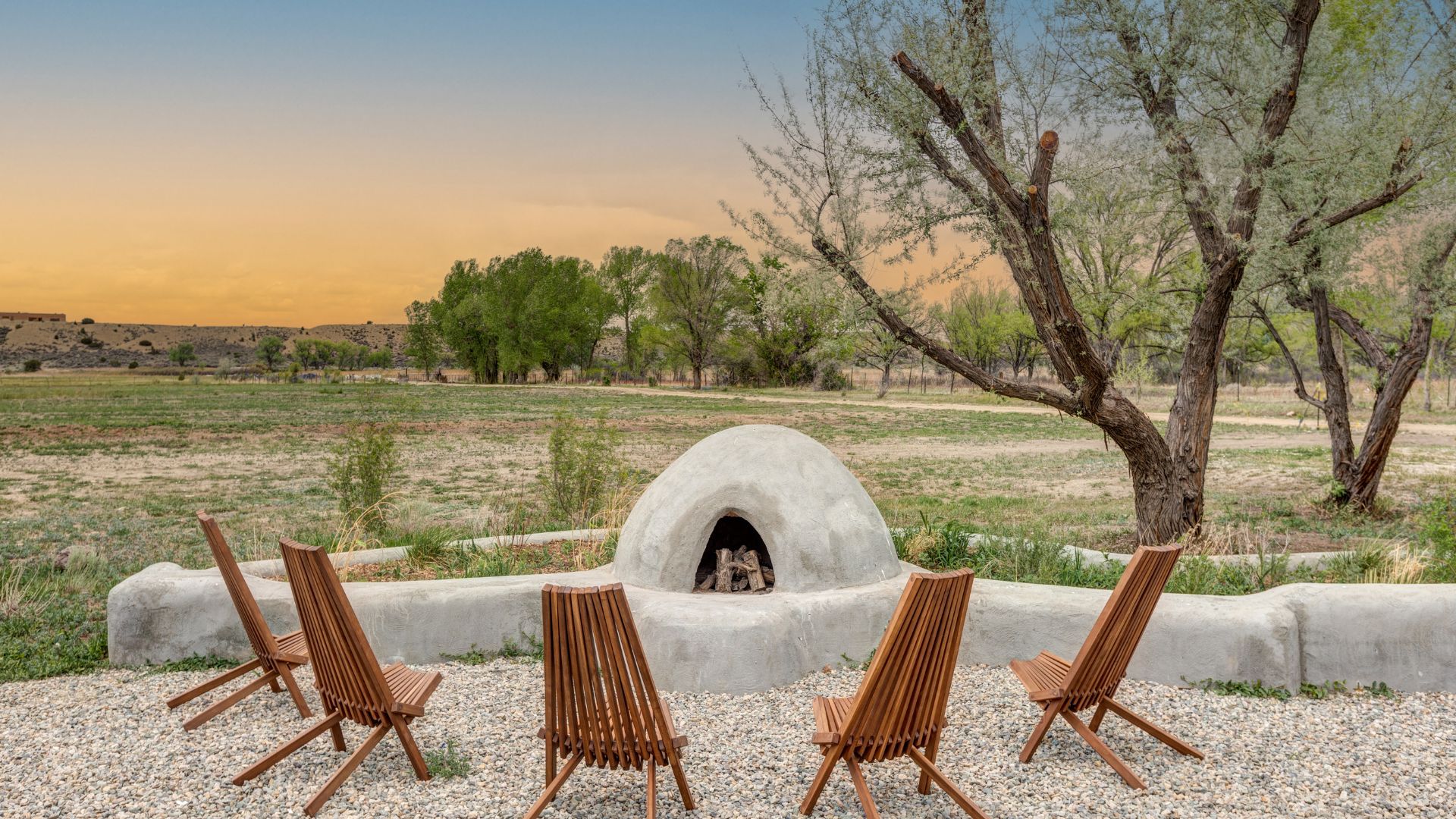 Orange and blue sunset in sky behind outdoor fireplace with five chairs